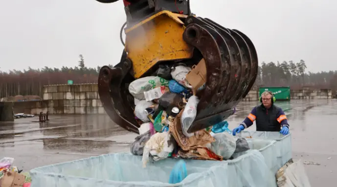 Bilden visar en gripklo som släpper avfall över en sorteringsbinge. En medarbetare övervakar arbetet.